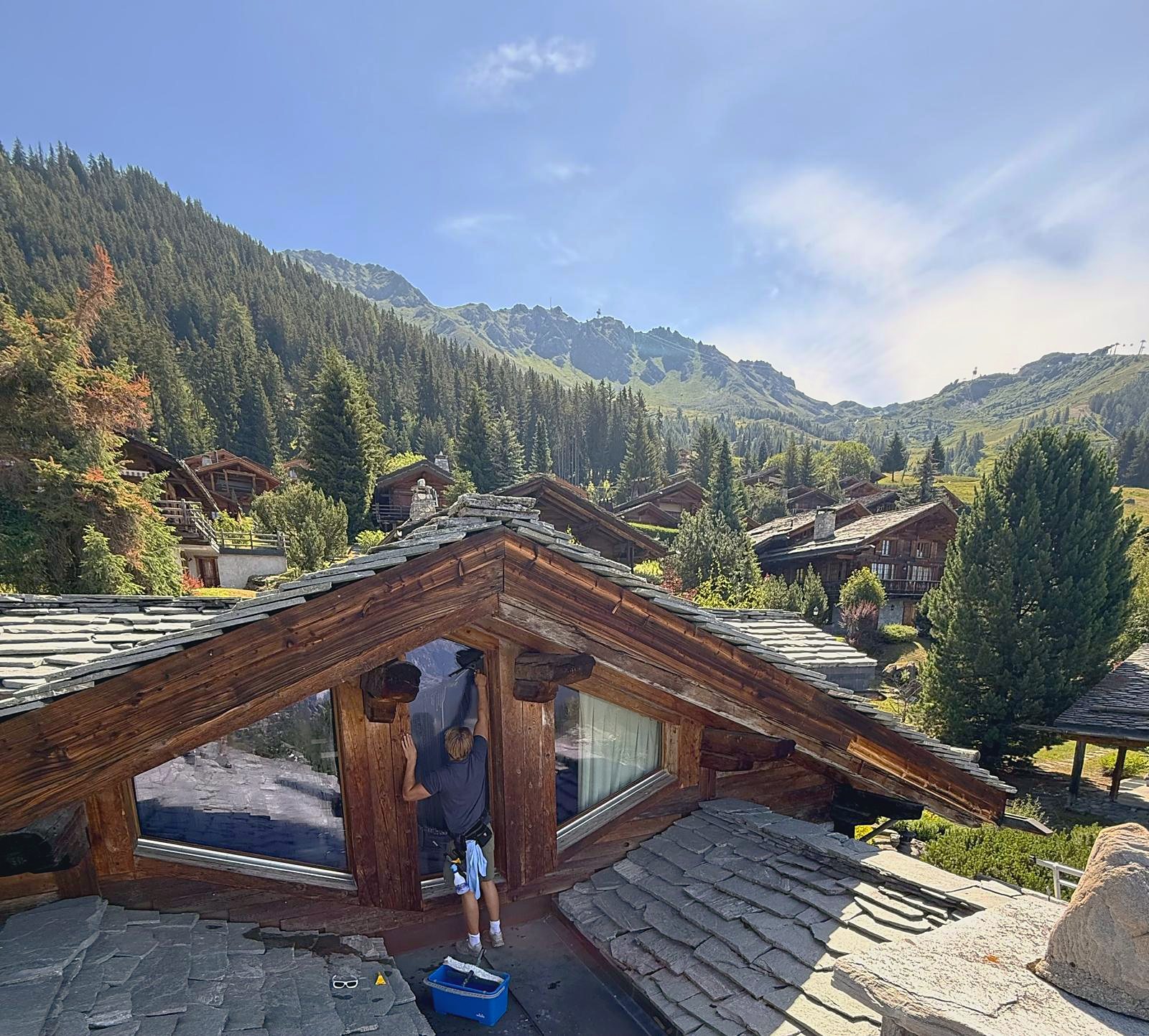 Cleaning a dormer window on a stone-tiled chalet roof above Verbier, looking out over the chalets and mountains of the Val de Bagnes.