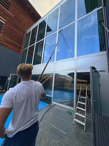 Water-fed pole cleaning a two-storey glazed facade beside a pool.